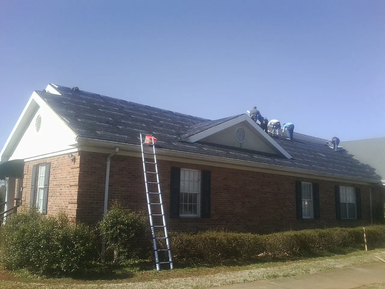 Crew tearing shingles off a brick ranch roof with a ladder set — Emergency Roof Repair work by our crew in Bellona