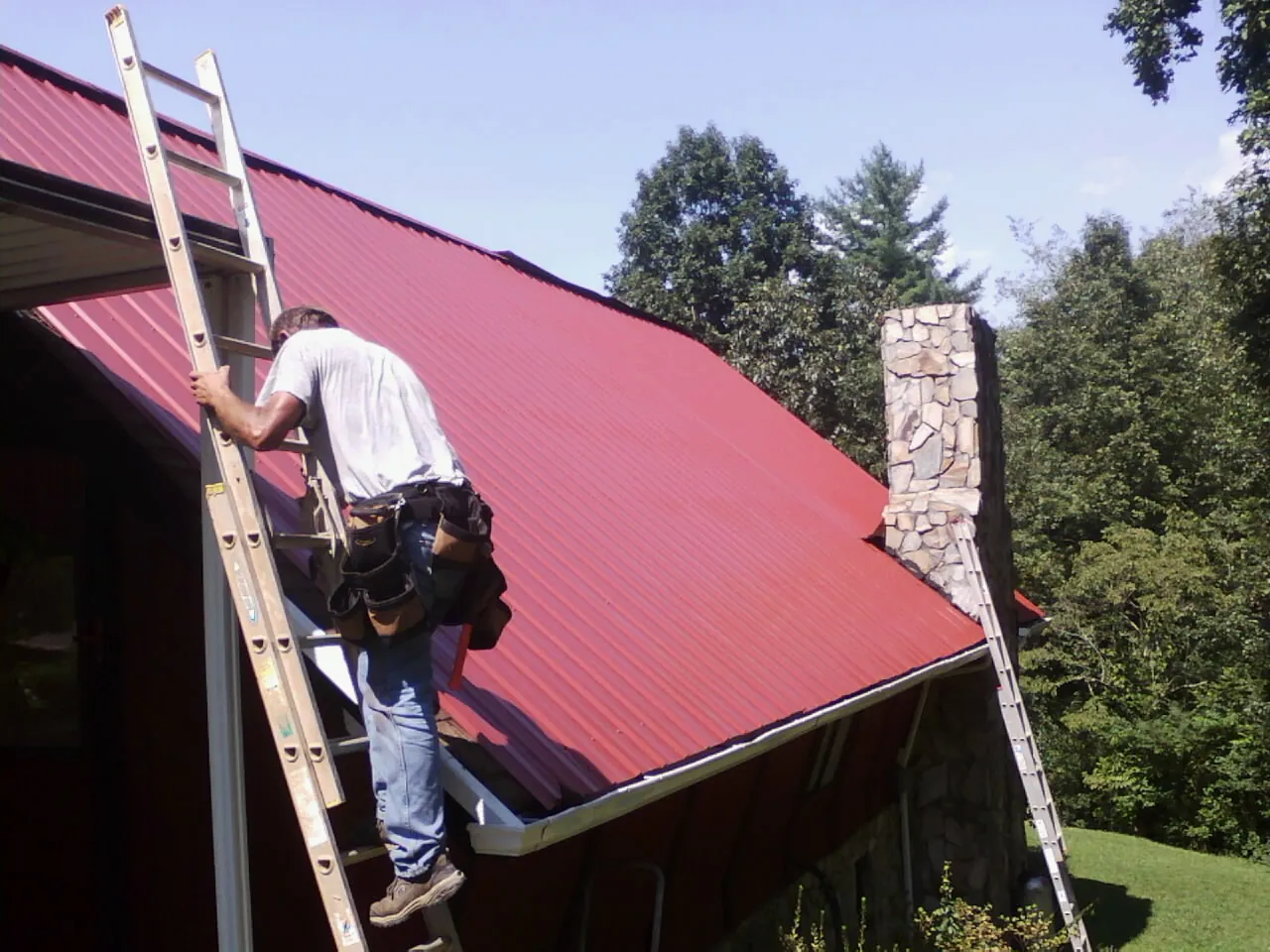 Roofer on a ladder beside a red metal roof and stone chimney — Soffit & Fascia Repair by Roofing Brothers in Bellona